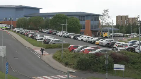 A wide shot of the busy car park at University Hospital, East Kilbride - a grey-clad building with red brick, looking onto the ambulance entrance of the A&E department 