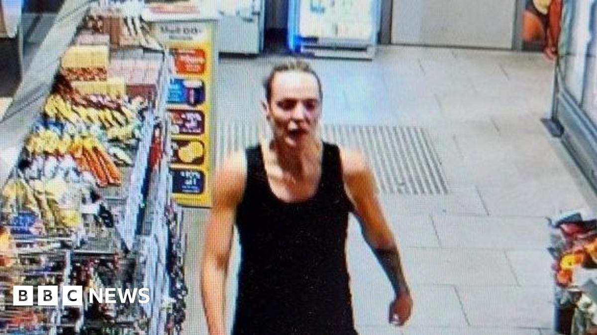 CCTV still of Rachel Booth with blonde hair tied back tightly. She walks past shelves of snacks to the garage till. She is wearing a sleveless black top.