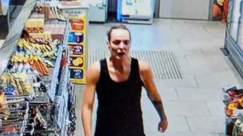 Cheshire Police CCTV still of Rachel Booth with blonde hair tied back tightly. She walks past shelves of snacks to the garage till. She is wearing a sleveless black top.