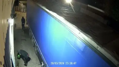 Metropolitan Police A man pours a green jerry can while another films him on a phone at the side of a lorry - all captured on security camera