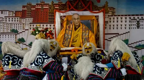 Reuters Tibetans in yellow masks and with white fluffy headdresses on, in colourful long-sleeve outfits perform a traditional mask dance in front of a cut-out of the Dalai Lama wearing all orange and yellow atop a throne