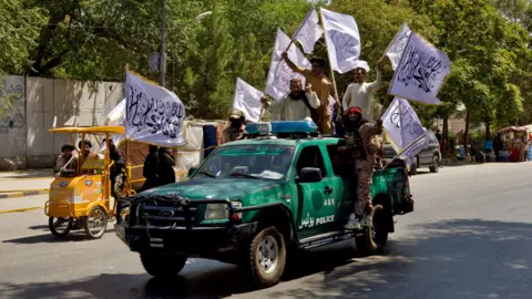 Reuters A battered green police vehicle is driving down a tree-lined street, with five men hanging off the sides waving white flags with Arabic text on them. The men are cheering and wearing elements of military dress, and two of them have large guns strapped to their chests. 