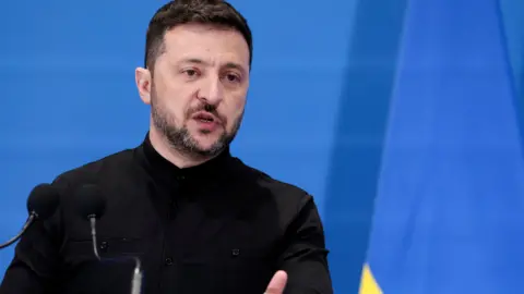 Reuters A close up of Volodymyr Zelensky speaking during a conference in Italy. He's wearing a black suit and is standing in front of a blue backdrop.