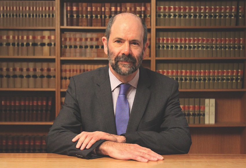 A man in a dark grey suit and blue tie sits with his hands resting on a wooden desk in front of a large bookshelf filled with academic texts.