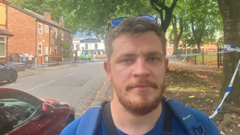Richard Stead/BBC A man with a gingerish beard and sunglasses on his head stands wearing a backpack next to a road. Behind him the road has been blocked off with police tape.