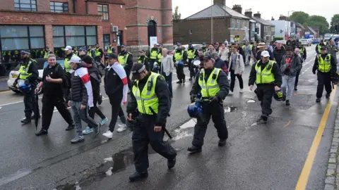 PA Media Police are walking alongside protesters on a road near the Bell Hotel in Epping. Some people can be seen with England flags over their shoulders