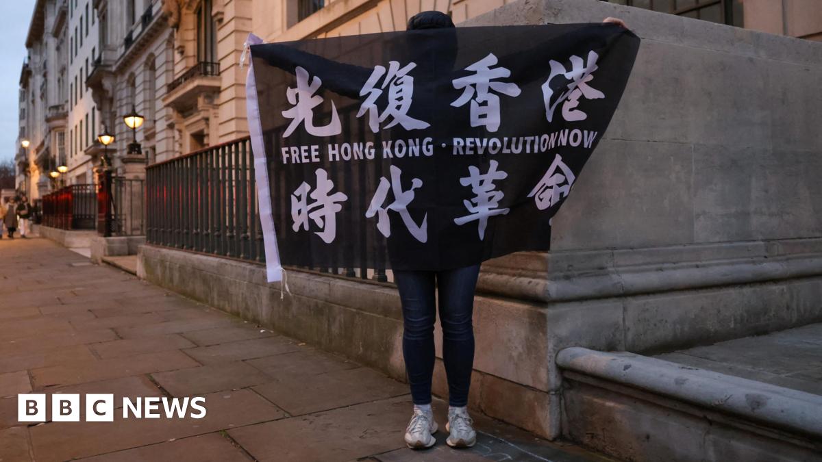 A demonstrator holds a black flag with white text that reads "Free Hong Kong - Revolution now" during a protest supporting the 45 pro-democracy activists sentenced to jail by Hong Kong's High Court. It is evening or early morning, and the demonstrator is on the street. The demonstrator's face his hidden.