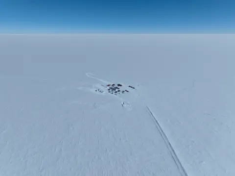 PNRA/IPEV A small group of tents and temporary shelters in the middle of the bright white Antarctic ice sheet. A trail of disturbed snow leads from the camp suggesting transport route. The sky is bright blue.