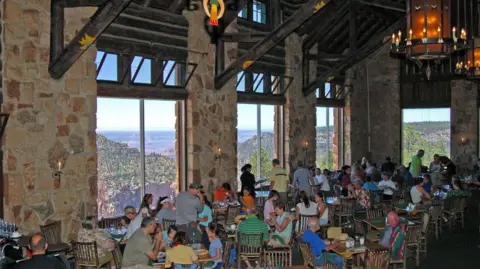 National Park Service Dining room with high ceilings and windows overlooking Grand Canyon, where people sit to eat or stand, during the day
