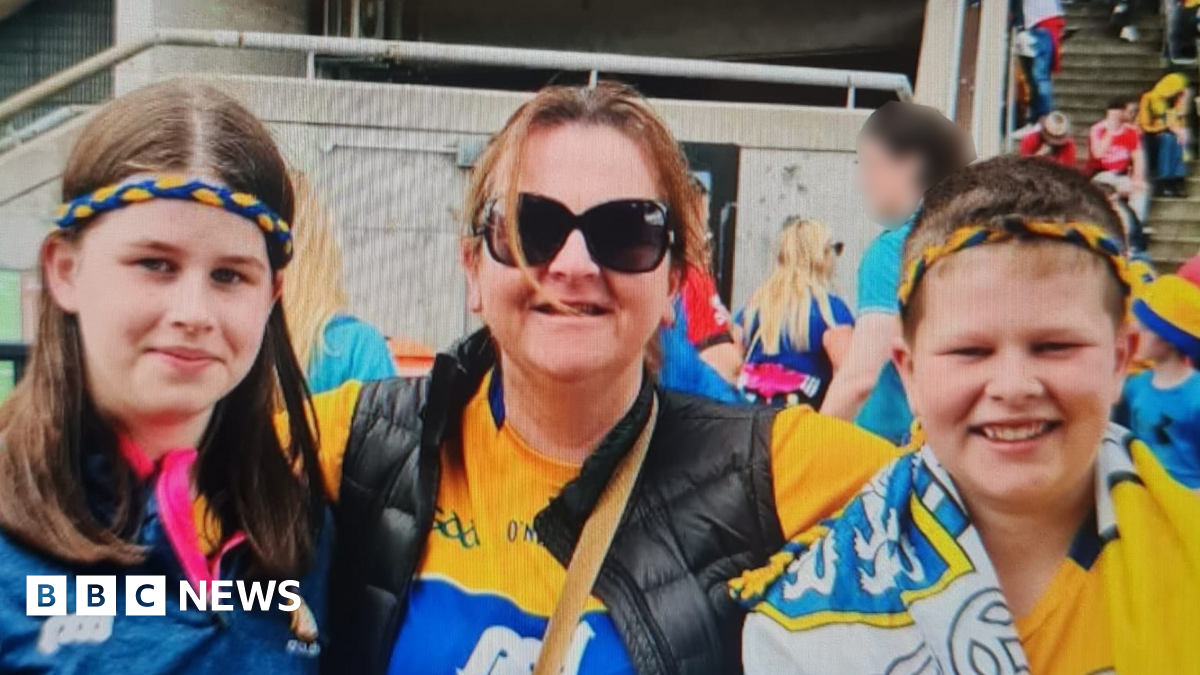A woman stands in the centre. A girl is on one side and a boy on the other side. A lot of other people are behind them. The woman is wearing sunglasses and a GAA jersey. The boy and girl have blue and yellow bands on their heads. The boy has a flag draped over his neck. They're all smiling.