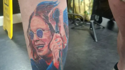 Simon Humphreys A tattoo of a smiling Ozzy Osbourne. In the image he is waring blue round glasses and a blue jacket and red top. Ozzy is also holding a microphone and has a tattoo on his fingers. 