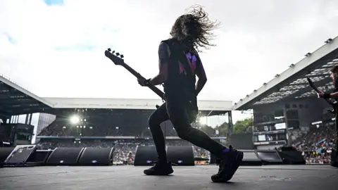 AFP via Getty Images A man with a guitar and long hair plays on a stage, in front of a large crowd.