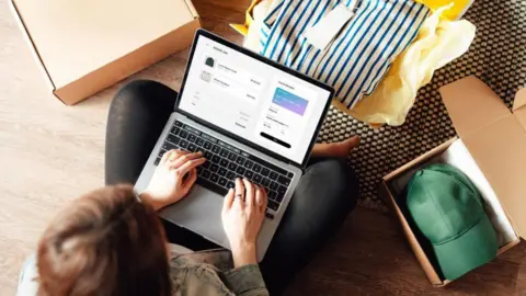 Getty Images Overhead shot of person on a laptop computer with a shopping website on the screen. A green cap is in a open box next to them.
