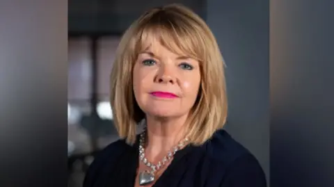 Warwickshire County Council Monica Fogarty looks at the camera. She has blonde hair and is wearing a silver heart shaped necklace. 