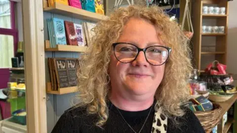 BBC Cherylee Barker smiles at the camera. She is stood in a cafe which shelves and chocolate bars in the background,