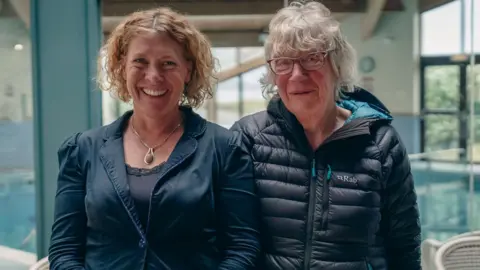 BBC News Two women stand facing the camera smiling. The women on the left has short curly ginger hair and is wearing a navy jacket. The woman on the right has short grey hair, glasses and a dark coat. 
