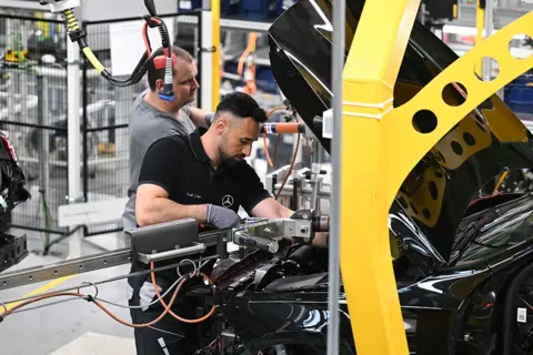 Getty Images A factory worker peers under the bonnet of a black car and holds a mechanical arm in place as he works