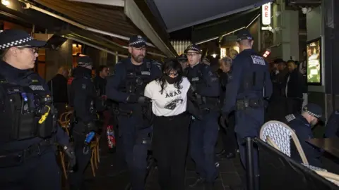 JOSH STANYER/EPA/Shutterstock Police take away an anti-Israeli protester with a black face mask, long dark hair and a white top from the restaurant where more police officers and others can be seen