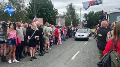 Richard Knights/BBC Scores of people standing on the road, many with St George's flags. A car is driving through the middle.