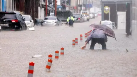 Reuters A man, hunched over and holding a polka-dotted umbrella, makes his way through a flooded street, caused by torrential rain, in Gwangju, South Korea on 17 July, 2025. A row of vehicles are parked on the left of the street. The floodwaters have submerged parts of the vehicles' wheels. 