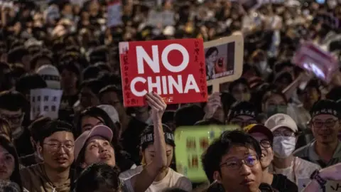 Getty Images At a night-time rally, a woman in a pink cap holds up a red and white sign that says in English "No China". Surrounding her are other protesters holding up other signs as well.