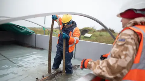 Orkney Islands Council A man standing in a water tank wearing high vis clothing, moving wooden planks in the water.