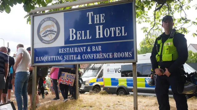 A police officer in black and a yellow high-vis jacket stands next to a blue sign reading "The Bell Hotel - restaurant and bar banqueting suite and conference rooms". There are a group of people huddled in the background and a police van in the background.