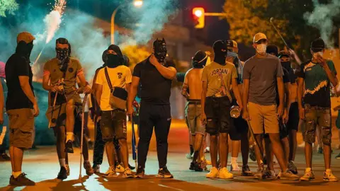 Getty Images A group of around a dozen men, mostly wearing shorts, t-shirts and trainers, wear masks and balaclavas as they stand around on a street at night time. Some hold baseball bats, sticks and chains, while a flare is lit in the background