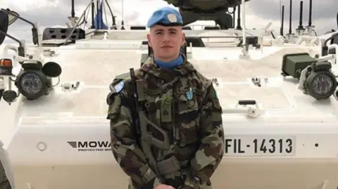 Irish Defence Forces Irish soldier Seán Rooney, wearing camouflage clothes and a blue beret. He is holding a gun and standing in front of an armoured vehicle.  