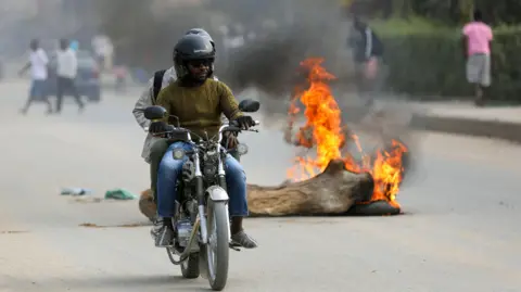 EPA People on a motorbike drive past a tree stump set on fire in Luanda on Monday.