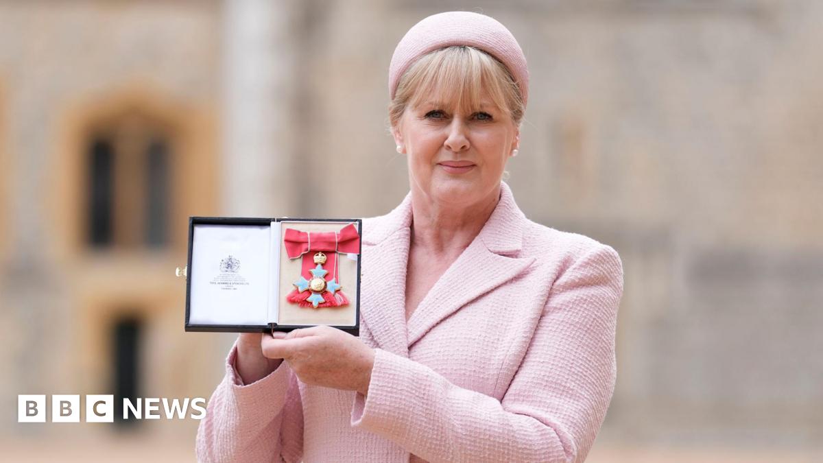 A mid-shot of Sarah Lancashire holding her CBE medal. She is wearing a pink beret and suit.