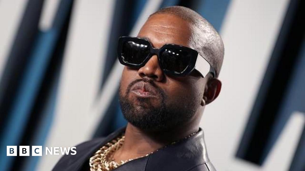 Kanye West with sunglasses on posing for a picture. He is also wearing a gold chain.
