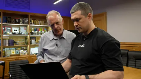 BBC/Xavier Vanpevenaege Two men stand looking at a laptop, one in the foreground in a black T-shirt, the other in a grey shirt
