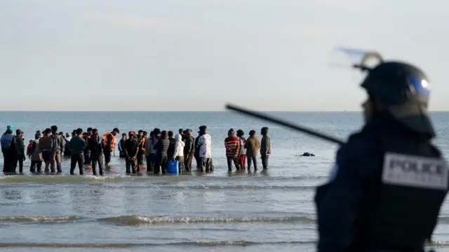 People thought to be migrants wade through the sea to board a small boat leaving the beach at Gravelines, France. A policeman is seen in the foreground.