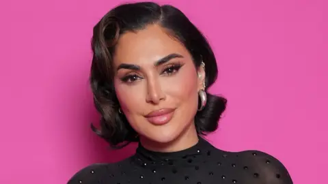 Getty Images Huda Kattan in a sequin-studded black sheer top smiling against a pink backdrop