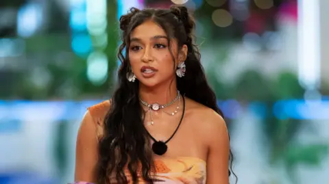 Ben Symons/Peacock via Getty Images Cierra Ortega on Love Island USA. Cierra has long, wavy dark hair and brown eyes. She wears a patterned one-shouldered orange dress and a microphone around her neck. 