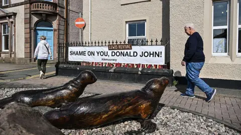 Getty Images A street in Stornoway. Outside a cream-coloured harled building, a banner saying 'Shame on you, Donald John' is attached to railings. A woman and man are walking past, one behind the other. There are two bronze sculptures of seals on the ground in front of them.