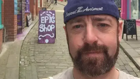 Facebook Ben Gunnery has a goatee beard and is wearing a purple beanie hat.  He's taking a selfie photo in a narrow street full of shops.