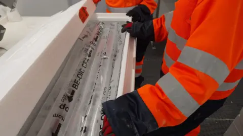 BBC News A person wearing orange and black protective clothes and gloves has their hands over long tubes of ice covered in plastic wrapping. The tubes are inside a large box.