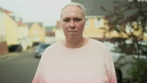 A woman with tied back blonde hair looks at the camera. She is not smiling. She is wearing a pale pink jumper and is standing in a residential street, which is blurred