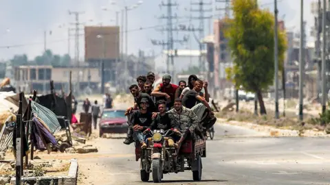 AFP Men ride on a motorised cart along Salah al-Din road in Deir al-Balah, as smoke billows in the background, in the central Gaza Strip (21 July 2025)