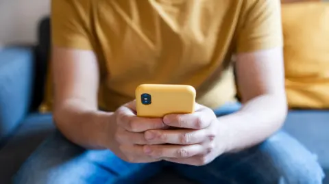 Getty Images A man sitting on a sofa wearing a yellow t-shirt and jeans, holding a yellow smartphone in his hands in front of him.
