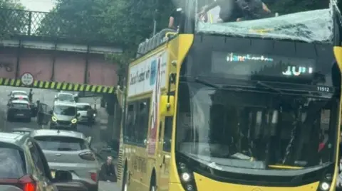 X People are standing on the top deck of a yellow bus of which the top has been pulled off. The front windscreen has been smashed. 