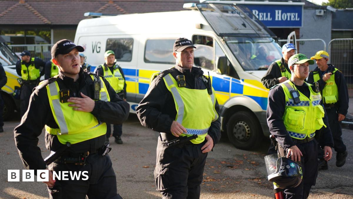 A group of police officers in fluorescent tabards speaking into talkback devices and holding riot helmets - there is a large police van in the background at the entrance to a building with a Bell Hotel sign