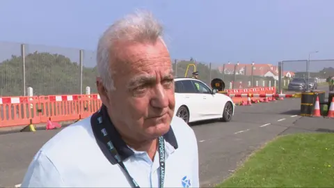 Gavin Scott looking slightly off camera. He has grey hair which is sticking up at the front. He is wearing a white polo shirt with a dark collar and a Saltire logo on the upper left chest. He is wearing a green lanyard around his neck with white writing on it.
