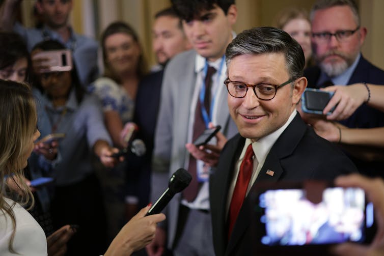 A white man with glasses, dark hair and a dark suit with a white shirt and red tie smiles and appears to speak into a microphone as people surround him.