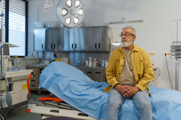 An older bearded white man in a yellow shirt sits on a hospital bed in an exam room
