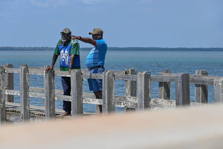 Two men stand on a wharf, one pointing into the distance