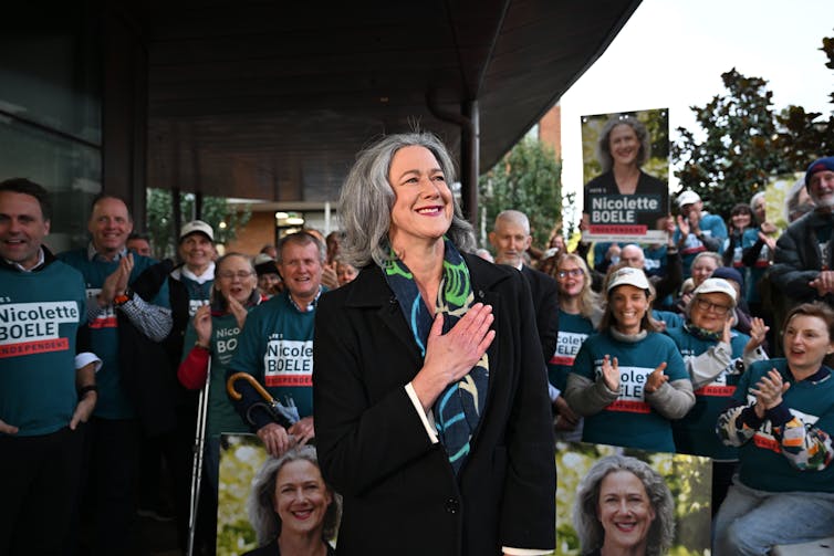 A woman with grey hair smiles in front of a large crowd holding signs