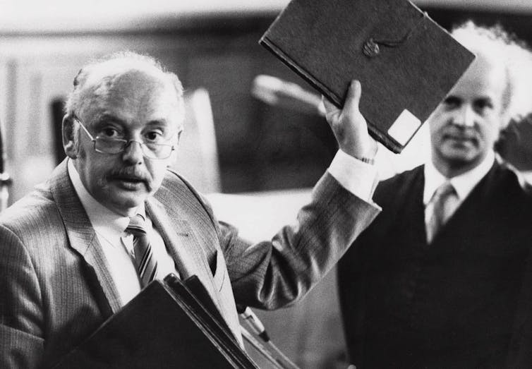 A man with glasses and a moustache holds up a black folder.
